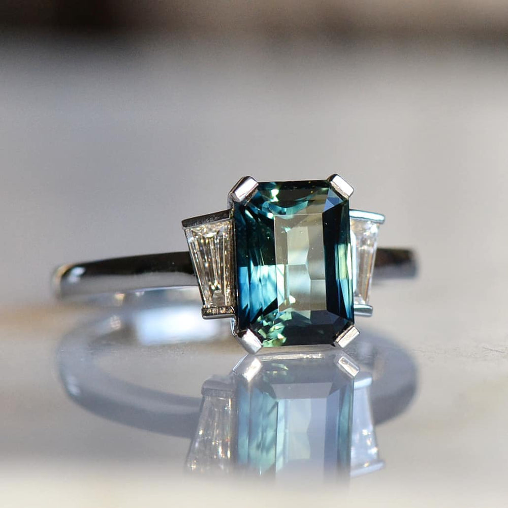 A detailed Emerald cut blue sapphire diamond ring with its reflection on a shiny surface