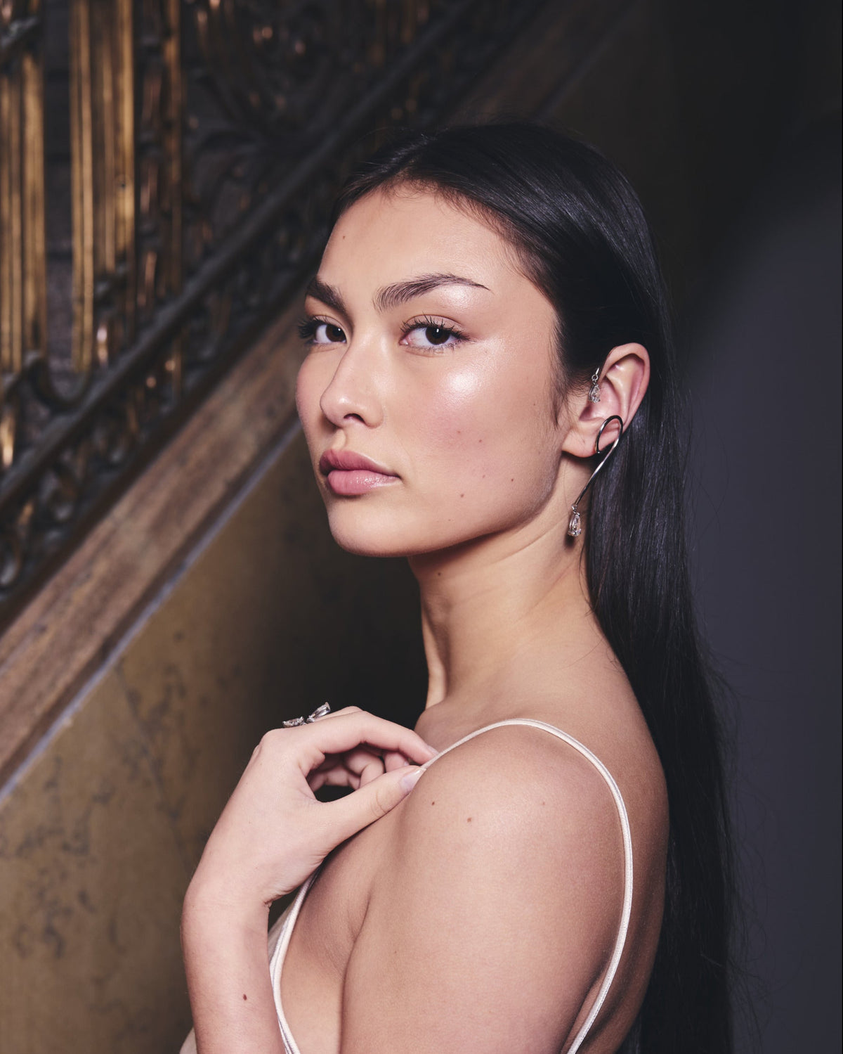 Woman in a white dress wearing white gold earring and rings standing against a dark wall with decorative elements.