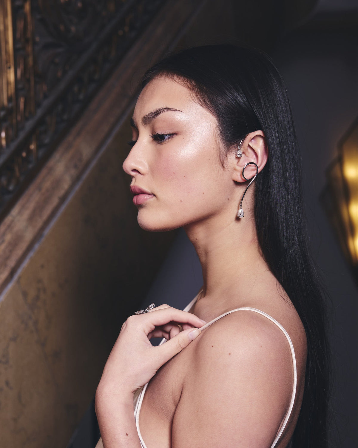 Woman in a white dress wearing white gold earrings and rings standing in front of brass and marble staircase 