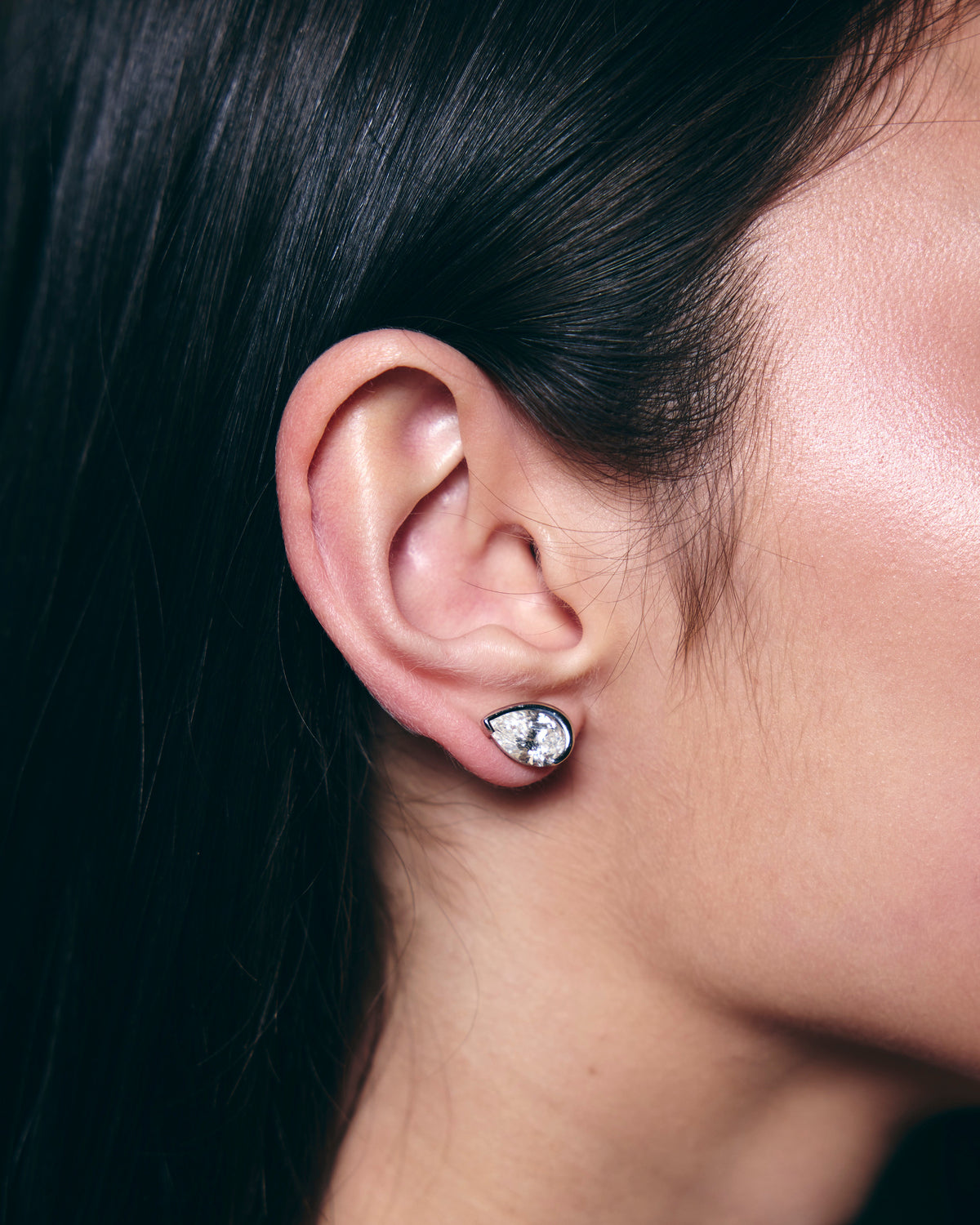 A woman with black hair showing a close up of her ear wearing a pear cut diamond earring stud. 