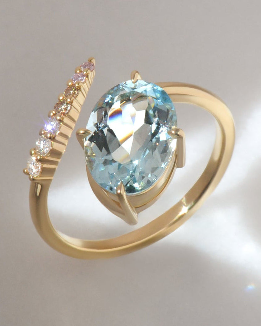 18ct gold ring featuring a large oval aquamarine centre stone, and a trail of small white diamonds, champagne diamonds, and faint pink diamonds on a curved open band, photographed on a light neutral background with bright sunlight and shadows.