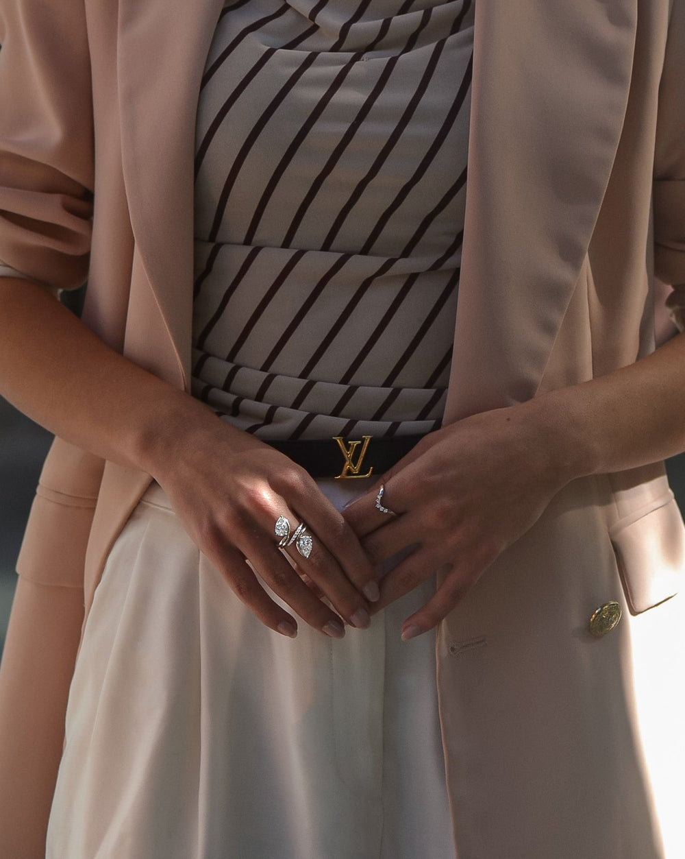 Person wearing a beige trench coat with a patterned bag and diamond rings.