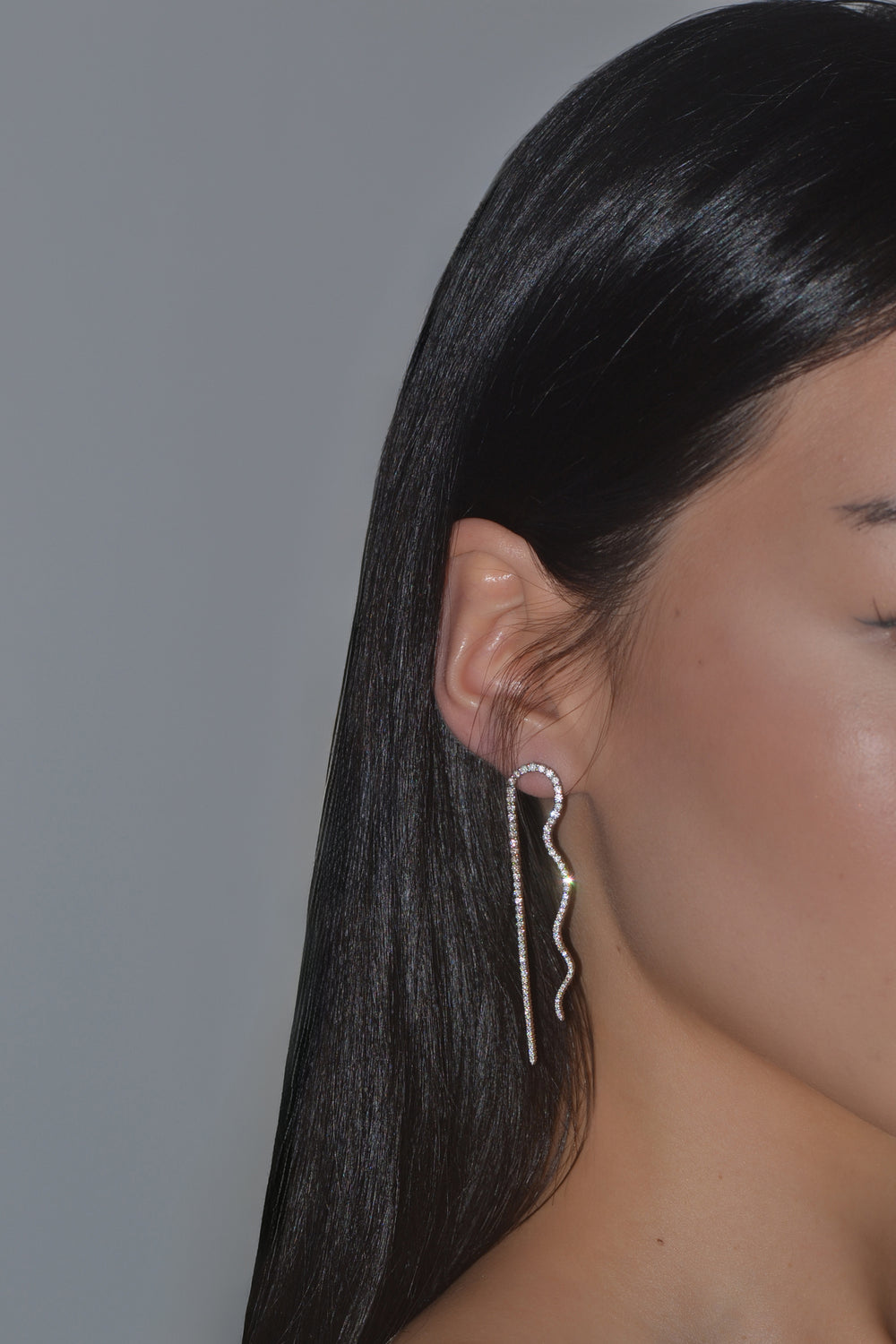 A close-up side profile of a woman with long black hair wearing a long, wavy diamond earring, photographed on a plain gray background.