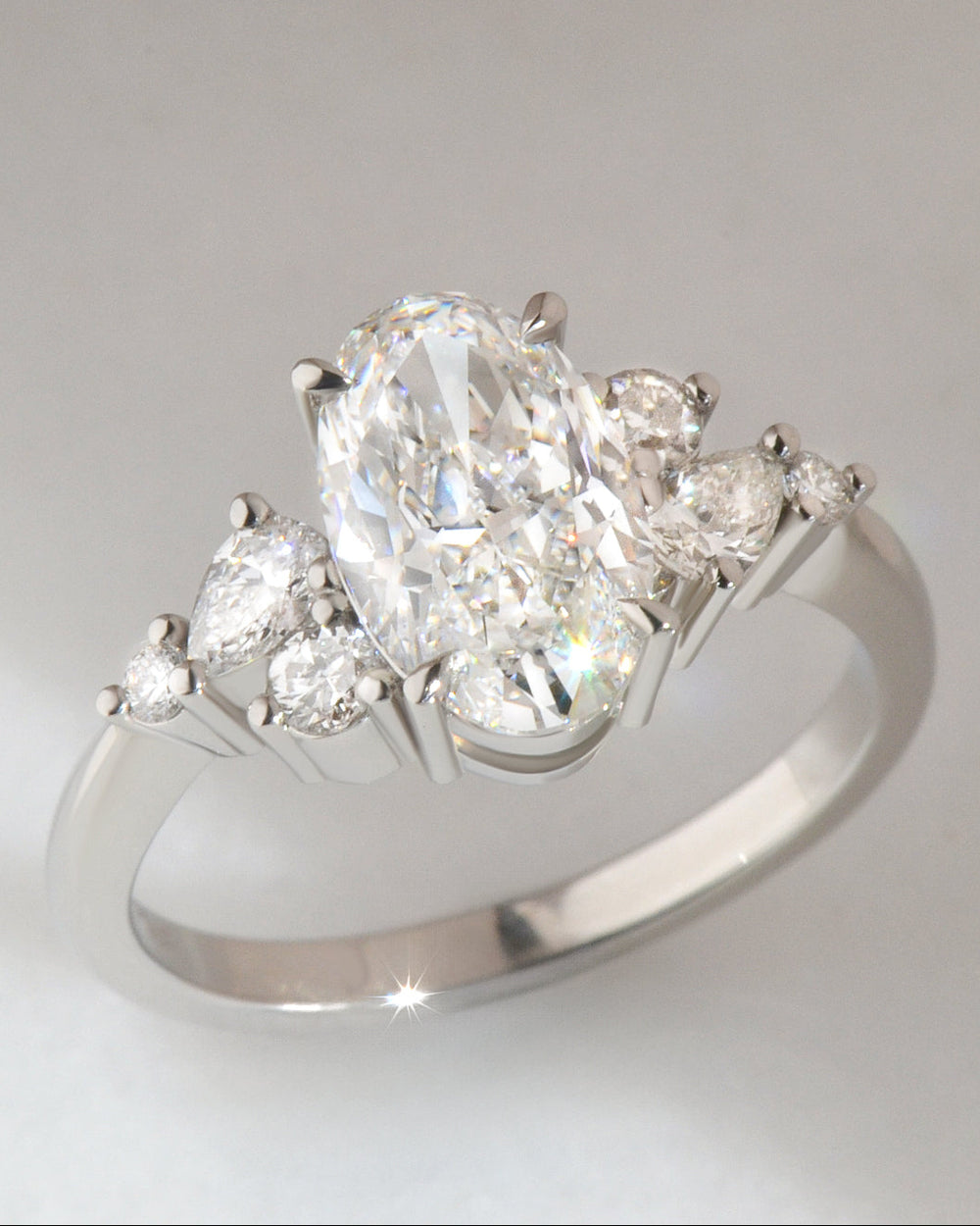 Diamond ring with a central oval cut  diamond and smaller round and pear cut diamonds set in platinum, on a light gray background