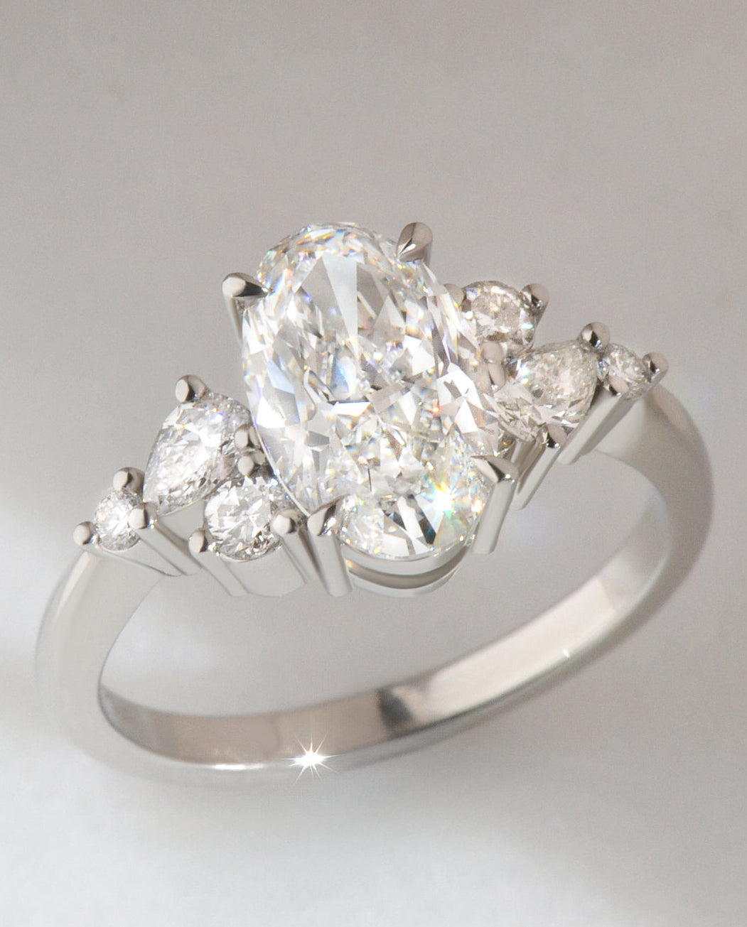 Diamond ring with a central oval cut  diamond and smaller round and pear cut diamonds set in platinum, on a light gray background