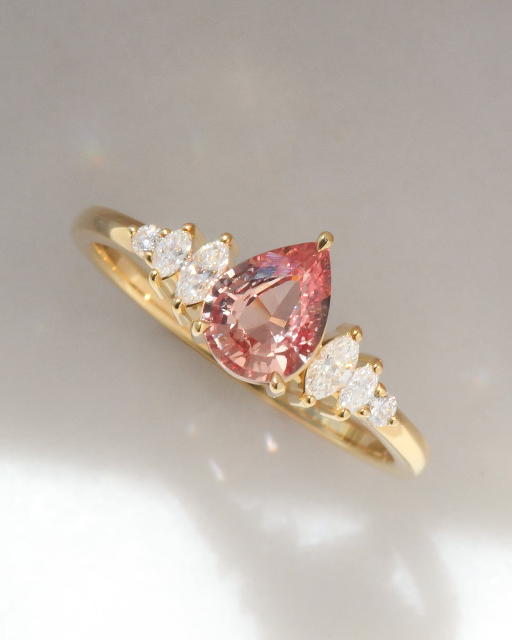 18ct yellow gold ring featuring a pear-cut Padparadscha sapphire centre stone, flanked by marquise cut diamonds on either side sides, photographed against a light grey background with a soft shadow.