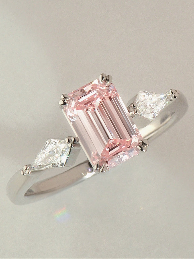 Emerald pink diamond ring with two kite cut diamonds set in platinum on a light grey background