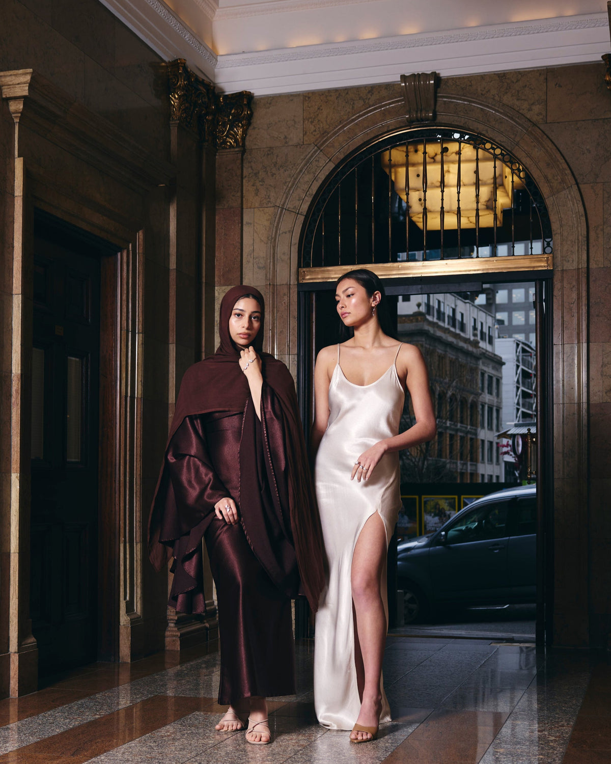 Two women in elegant outfits standing in a grand hallway with a cityscape view.