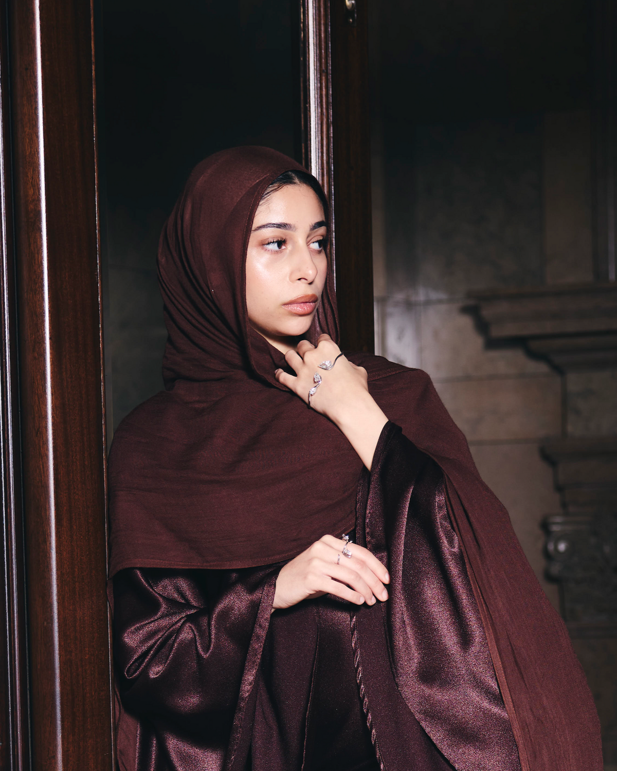 Woman wearing a dark brown hijab and dress standing against a stone wall.