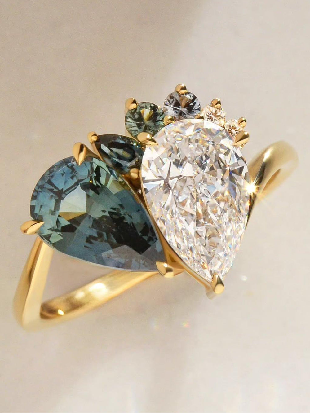 18ct yellow gold ring with a pear cut diamond pear cut sapphire Toi et Moi ring with small sapphires and diamonds surrounding the setting, on a light background.